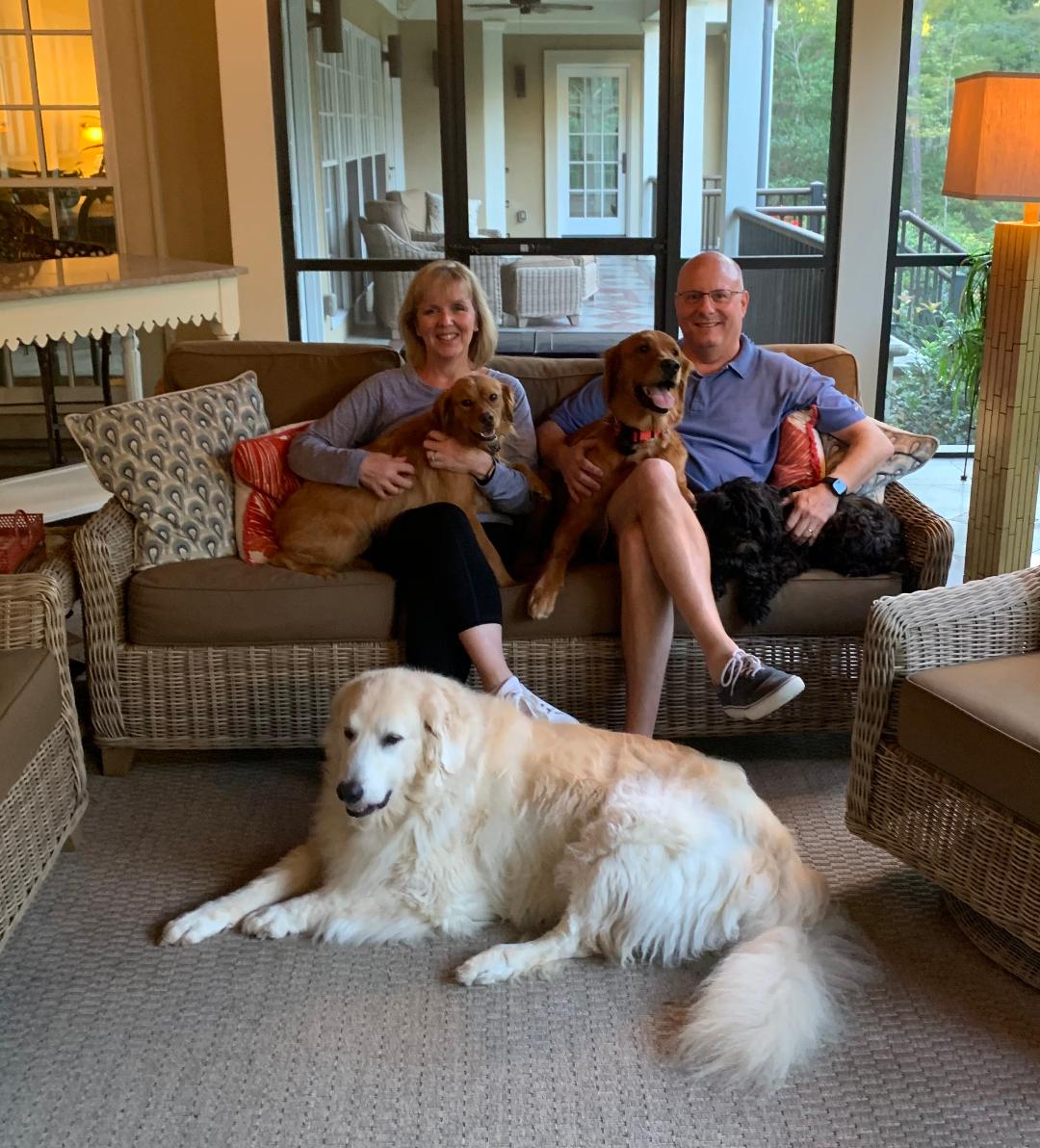 Couple sitting on couch holding two golden retrievers and one golden retriever laying in front of them